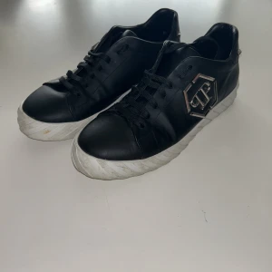 Philipp Plein svarta sneakers med detaljer - Svarta sneakers från Philipp Plein med vit platt sula och snörning. Sidan pryds av en stor metall-logga och bak finns coola döskalle-detaljer i silver. Skorna är gjorda i skinn och har en rund tå. Levereras med originalkartong och dustbag.