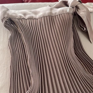 Plisserad beige omlottkjol med knytband - Snygg plisserad omlottkjol i beige med glansig finish och breda knytband i midjan. Kjolen har ett elegant fall och är tillverkad i ett satinliknande material som ger en lyxig känsla. Perfekt för dig som gillar stilrena och trendiga plagg med extra detalj.