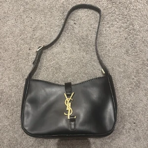 Svart axelväska från Yves Saint Laurent - Snygg svart axelväska från Yves Saint Laurent med klassisk YSL-logga i guld på framsidan. Väskan är i slät skinn och har justerbar axelrem samt dragkedja upptill. Perfekt storlek för det viktigaste och ger en lyxig vibe till din outfit.