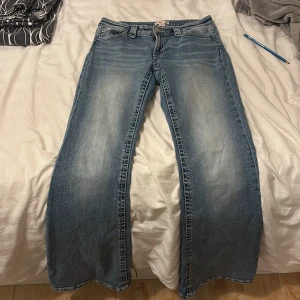 Blå bootcut jeans från Gina Tricot - Snygga blå jeans från Gina Tricot i bootcut-modell med ljus tvätt och slitningar framtill. Jeansen har klassiska fickor och coola detaljer med nitar på bakfickorna. Perfekt för dig som gillar en avslappnad och trendig look.