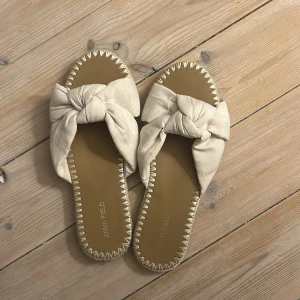 Beige sandaler med rosett från Anna Field - Snygga beige sandaler från Anna Field med breda tygband i omlott och stor rosettdetalj upptill. Platt sula med dekorativa sömmar runt kanten och öppen tå. Perfekta för sommardagar och lätta att matcha till olika outfits.