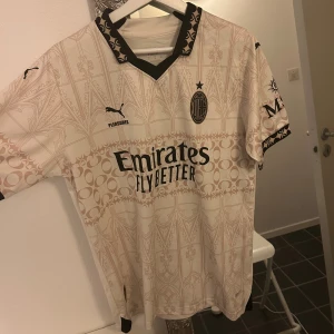 AC Milan Rafa Leão fotbollströja - Säljer en AC Milan fotbollströja med Rafa Leão och nummer 10 på ryggen. Tröjan är ljusbeige med detaljerat mönster, svarta tryck och loggor, samt svarta mönstrade muddar vid krage och ärmslut. Materialet är lätt och andas, perfekt för match eller träning.