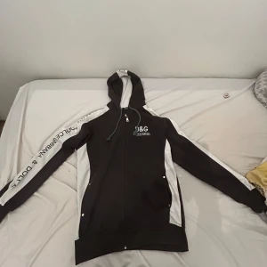 Svart och vit D&G hoodie XL - Snygg svart och vit hoodie från Dolce & Gabbana i storlek XL. Tröjan har dragkedja, huva med snörning och logga broderad på bröstet samt tryck längs ärmarna. Perfekt för dig som gillar streetstyle och vill sticka ut. Passar även X samt M. Fick den av min morbror och vill inte ha den 