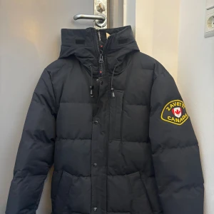 Svart pufferjacka från Zavetti Canada - Svart pufferjacka från Zavetti Canada med huva och snyggt broderat märke på ärmen. Jackan har dragkedja, knappar och snörning vid halsen. Flera praktiska fickor framtill och quiltad design som ger en cool look. Perfekt för kalla dagar. Kan gå ner i pris vid snabb affär 