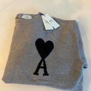 Grå stickad tröja från Ami Paris - Snygg grå stickad tröja från Ami Paris med svart hjärta och A-logga på bröstet. Klassisk rund halsringning och ribbade muddar. Perfekt för dig som gillar stilrena detaljer och vill ha något som sticker ut lite extra.