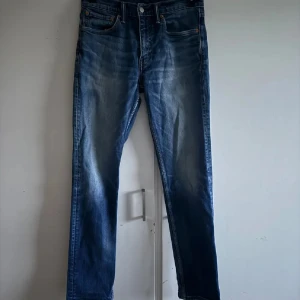 Levi's 511 blå jeans W31 L32 - Säljer ett par klassiska Levi's 511 jeans i blå tvätt i storlek 31/32. Modellen är slim och har raka ben, fem fickor och orangea sömmar. Jeansen är tillverkade i bomull och har en ikonisk läderpatch bak i midjan. Lite slitet vid knäet (bild 4) och där bak (bild 5) men syns knappt