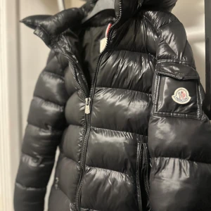 Svart Moncler pufferjacka med huva - Säljer en svart pufferjacka från Moncler med ikonisk logga på ärmen och stor huva. Jackan har blanka paneler, dragkedja framtill och en ficka på ärmen. Insidan har ett coolt serietidningsmönster på etiketten. Perfekt för kalla dagar.