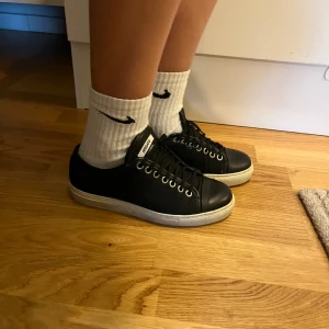 Svarta sneakers från Axel Arigato - Svarta sneakers från Axel Arigato med rund tå och platt vit sula. Skorna har snörning med svarta skosnören och silverfärgade öljetter. Tillverkade i skinn med en stilren och minimalistisk design, perfekt för dig som gillar cleana och klassiska sneakers.
