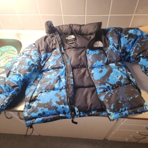 Blå camo pufferjacka The North Face XS - Säljer en fet pufferjacka från The North Face i storlek XS. Jackan har blått och svart camomönster, hög krage och dragkedja framtill. Perfekt för kalla dagar och riktigt snygg streetstil. Logga på bröstet och insidan. Fylld med dun för extra värme.