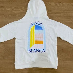 Casablanca hoodie (helt ny+gratis frakt) - Vit hoodie från Casablanca med färgglad grafisk print på både bröstet och ryggen. Trycket har blå, gul och orange toner och texten 'Casa Blanca'. Hoodien har huva och känguruficka framtill. Perfekt för en chill och stilren look.