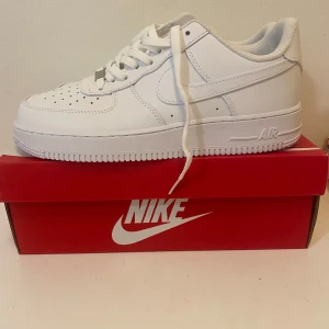 Nike Air Force 1 vita sneakers - Nike Air Force 1 sneakers i helvit färg med klassisk låg siluett. Skorna har snörning, rund tå och platt sula. Tillverkade i skinn med perforerade detaljer på tån för extra ventilation. Snygg och tidlös modell som passar till allt.