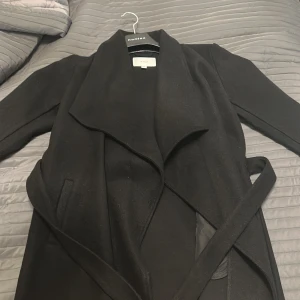 Svart kappa med bälte från Vila - Stilren svart kappa från Vila med bred krage och knytskärp i midjan. Jackan har långa ärmar, diskreta fickor och en snygg omlott-look. Perfekt för dig som gillar en klassisk och clean stil.