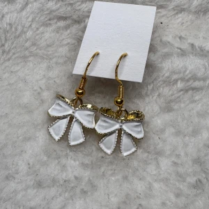 Örhängen med vita rosetter - Supersöta örhängen med vita rosetter och guldfärgade detaljer. Hängande modell som ger en gullig och lekfull vibe, perfekta för att lyfta vilken outfit som helst. 