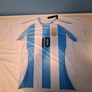 Argentina Messi #10 fotbollströja - Säljer en klassisk Argentina fotbollströja med Messi och nummer 10 på ryggen. Tröjan har blå och vita vertikala ränder, korta ärmar och AFA-emblemet med tre stjärnor på bröstet. Tillverkad i lätt polyester, perfekt för match eller träning.