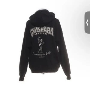 Svart Gymshark hoodie med tryck - Svart hoodie från Gymshark med stort tryck på ryggen och liten logga framtill. Oversized passform, huva och känguruficka. Tillverkad i 80% bomull och 20% återvunnen polyester. Perfekt för gymmet eller chill.