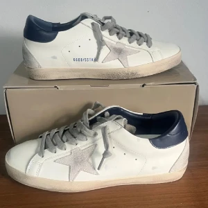 Golden Goose vita sneakers med stjärna - Snygga vita sneakers från Golden Goose med grå mockadetaljer och en ikonisk stjärna på sidan. Skorna har beige snörning, mörkblå häl och platt gummisula med fiskbensmönster. Perfekt för dig som gillar en cool och avslappnad stil.