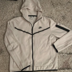 Vit nike tech - Nike hoodie i vitt med svarta detaljer och dragkedja hela vägen. Tröjan har huva, lång ärm och en snygg svart linje över bröstet samt logga på vänster sida. Tillverkad i mjukt material, perfekt för chill eller träning.