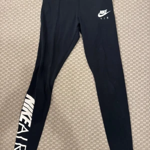 Svarta Nike Air leggings - Svarta leggings från Nike Air med vit logga på höften och stort tryck längs ena benet. Tight passform och stretchigt material som passar perfekt till träning eller chill. Snygg och sportig look med klassisk Nike-stil.