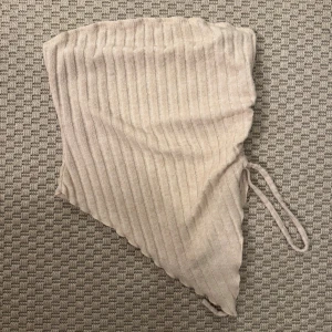 Beige ribbad kortkjol med snörning - Trendig beige kortkjol med ribbad struktur och snörning i sidan. Kjolen har asymmetrisk nederkant som ger en cool och modern look. Perfekt för dig som gillar stilrena och enkla plagg med en twist.