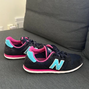 New Balance sneakers blå/rosa (nypris 899kr) - Snygga New Balance 373 sneakers i mörkblått med turkosa detaljer och rosa foder. Skorna har klassisk snörning, platt sula och rund tå. Materialmix av mocka och syntet med meshpartier för extra komfort. Perfekt för dig som gillar färgstarka och sportiga sneakers.