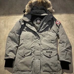 Grå parkas från Canada Goose med päls - Säljer en grå parkas från Canada Goose med ikonisk patch på ärmen, stor huva med päls och flera praktiska fickor. Jackan har dragkedja och knappar framtill, muddar vid ärmslut och en klassisk, rak passform. Perfekt för kalla vinterdagar. Hör av vid minsta fundering 💫