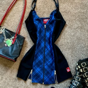 Blå och svart rutig klänning Living Dead Souls - Snygg kortklänning från Living Dead Souls med blått och svart rutigt mönster framtill, svarta sidor och justerbara axelband. Dragkedja hela vägen framtill och coola detaljer med dragkedjor vid nederkanten. Perfekt för dig som gillar alternativ stil.