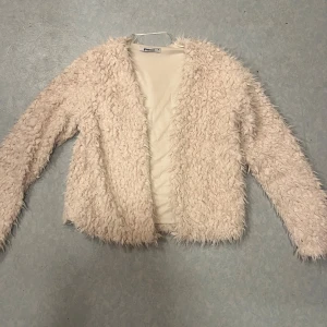 Beige pälsjacka från Gina Tricot - Fluffig och supermjuk beige pälsjacka från Gina Tricot. Jackan har lång ärm, öppen framtill och är fodrad på insidan. Perfekt för dig som vill ha en mysig och trendig look med lite extra attityd.