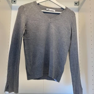 Grå v-ringad tröja från Zara - En stilren grå v-ringad tröja från Zara med lång ärm och ribbade muddar. Tröjan har en klassisk passform och är tillverkad i mjuk bomullsmix, perfekt för lager-på-lager under kyligare dagar.