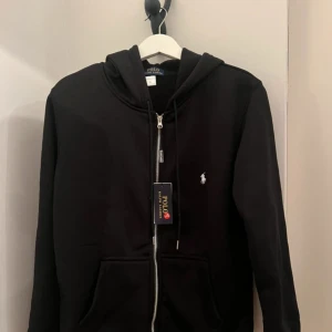 Svart zip från Polo Ralph Lauren - Svart hoodie från Polo Ralph Lauren med dragkedja, huva och snörning. Klassisk logga broderad på bröstet och två fickor framtill. Tillverkad i mjuk bomullsmix, perfekt för en chill och stilren look.