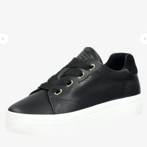 Svarta sneakers från Gant i skinn - Stilrena svarta sneakers från Gant med vit platt sula och gulddetaljer vid snörningen. Skorna är gjorda i skinn och har en klassisk rund tå. Gant-logga i guld på sidan, plösen och hälen. Perfekta för en clean och modern look.
