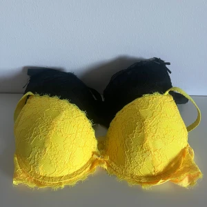 Gul och svart spetsig bh - Två snygga bh, en i knallgul och en i svart, båda med blommig spets och vadderade kupor. De har axelband och en liten rosett framtill för extra detalj. Perfekt för dig som gillar att sticka ut med färg eller köra klassiskt svart.