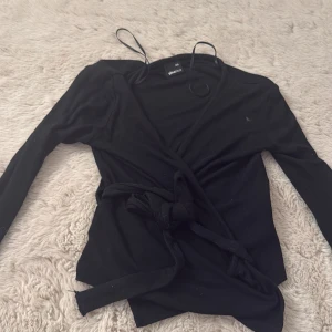 Svart omlottopp från Gina Tricot - Svart omlottopp från Gina Tricot i storlek XXS. Toppen har långa ärmar och knytband i midjan för en snygg siluett. Mjuk och stretchig bomullskvalitet som känns skön mot huden. Perfekt att styla med jeans eller kjol för en cool look.