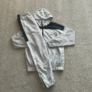 Lacoste grå och svart tracksuit - Säljer en stilren tracksuit från Lacoste i ljusgrått med svarta detaljer. Setet består av en jacka med huva, dragkedja och meshfoder samt matchande byxor med resår i midja och benslut. Ikoniska Lacoste-loggan syns på både jacka och byxa.