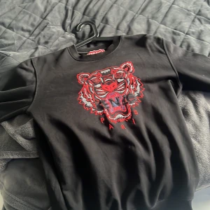 Svart Kenzo sweatshirt med tigerbrodyr - Svart sweatshirt från Kenzo med ikonisk tigerbrodyr i rött och grått framtill samt texten 'Paris'. Tröjan har rund halsringning och långa ärmar. Perfekt statement piece för dig som vill sticka ut med en cool och trendig look.