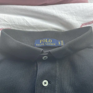 Svart pikétröja från Ralph Lauren M - Klassisk svart pikétröja från Ralph Lauren med röd broderad logga på bröstet. Tröjan har krage, två knappar och korta ärmar. Perfekt för en clean och stilren look. Tillverkad i mjuk bomull som känns skön mot huden.