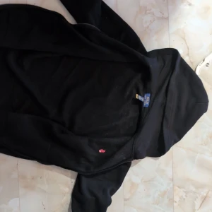 Svart hoodie från Polo Ralph Lauren - Svart hoodie från Polo Ralph Lauren med klassisk röd logga broderad på bröstet. Tröjan har huva, lång ärm, känguruficka och dragkedja framtill. Perfekt för dig som gillar stilren och enkel streetwear.