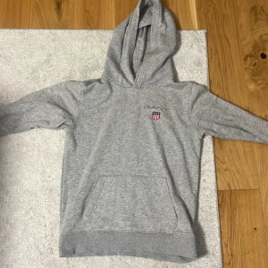 Grå hoodie från GANT med logga - Snygg grå hoodie från GANT med broderad logga och sköld på bröstet. Tröjan har huva, magficka och är tillverkad i mjuk bomullsmix. Perfekt för en chill och sportig look.