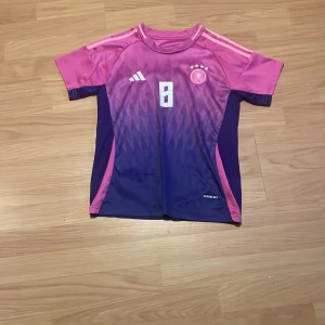 Adidas Tyskland Kroos #8 fotbollströja - Snygg Tyskland fotbollströja från Adidas med Kroos och nummer 8 på ryggen. Tröjan är kortärmad och har en cool gradient i rosa och mörklila med geometriskt mönster. Tillverkad i lätt polyester med Aeroready-teknologi för bästa komfort på planen.