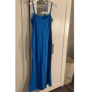 Blå långklänning med smala axelband - En stilren blå långklänning med smala axelband och rak passform. Klänningen har en enkel design utan mönster och är perfekt för varma dagar. Materialet känns mjukt och lätt mot huden.