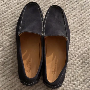 Svarta mockaloafers från Rizzo - Stilrena svarta loafers från Rizzo i mjuk mocka med kontrasterande vita sömmar. Skorna har platt sula med gummidetaljer undertill och klassisk rund tå. Perfekta för en clean och avslappnad look.