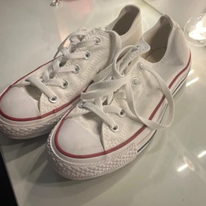 Vita Converse All Star sneakers strl 36 - Klassiska vita Converse All Star sneakers i canvas med röda och blå detaljer runt sulan. Lågt skaft, snörning framtill och rund tå. Perfekta för en clean och tidlös stil. Sula i gummi och ovandel i textil. Unisexmodell i storlek 36.