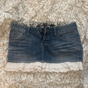 Jeanskjol med spetsvolang från Fishbone - Supersöt jeanskjol från Fishbone i klassisk blå denim med egensydd vit broderad spetsvolang längst ner. Kjolen har fem fickor, bälteshällor och knapp- och dragkedjestängning. Insidan har blommigt tygdetalj. Perfekt för dig som gillar en mix av denim och romantiska detaljer. Den är assnygga men tyvärr lite för liten så säljer den istället. Midjemått rakt över är 38cm, längd fram är 26 och längd bak är 30. 