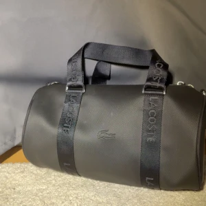 (ÄKTA) Svart duffelväska från Lacoste - Snygg svart duffelväska från Lacoste med cylinderformad design. Väskan har breda handtag med Lacoste-logga och är tillverkad i slitstark syntet. Perfekt för gymmet eller som weekendbag. Klassisk krokodil-logga på sidan för extra stilpoäng.