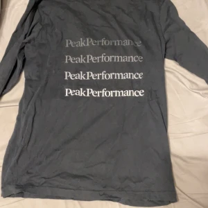 Svart långärmad Peak Performance-tröja - Svart långärmad tröja från Peak Performance med flera logotyper tryckta i vitt och grått på ryggen. Klassisk rund halsringning och mjukt bomullsmaterial som är skönt mot huden. Perfekt för dig som gillar stilrena och sportiga plagg.