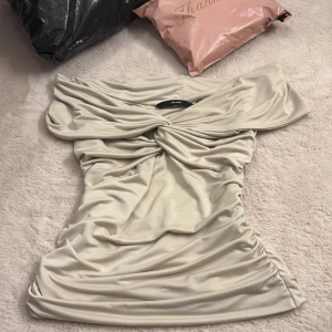 Topp Vero Moda - Superfin kort offshoulder topp från Vero Moda i champagnefärgad, glansig polyester. Snygg drapering och twistad detalj framtill ger en lyxig vibe. Skriv om ni har frågor. Gått ner i pris mycket eftersom den har en liten bronzer fläck💕