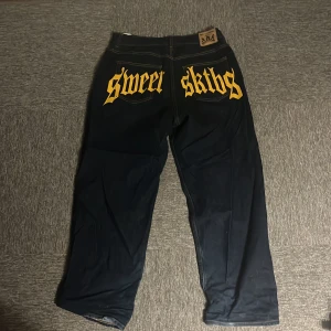 Sweet SKTBS mörkblåa jeans med tryck - Mörkblåa jeans från Sweet SKTBS i storlek M med stort gult tryck 'sweet sktbs' på bakfickorna. Klassisk femficksmodell med stora ben och kontrastsömmar. Jeansen har en baggy  passform och är tillverkade i bomull. Perfekta för dig som gillar streetwear och vill sticka ut.