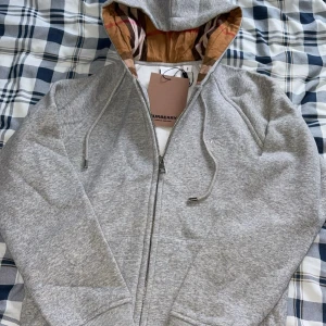 Grå hoodie från Burberry med dragkedja - Säljer en grå hoodie från Burberry med klassiskt rutigt foder i huvan och broderad logga på bröstet. Tröjan har hel dragkedja, snörning i huvan och ribbade muddar. Perfekt för dig som vill ha en stilren och lyxig vardagslook.