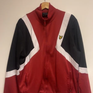 Röd och svart zip tröja  Lyle & Scott - Snygg zip tröja från Lyle & Scott i rött, svart och vitt. Tröjan har dragkedja framtill, ståkrage och två fickor. Materialet är polyester och bomull. Perfekt för dig som gillar sportig stil. Nypriset är ca 900 kr. Den är nästan helt oanvänd eftersom att storleken var förstor.