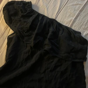 Svart oneshoulder blus  - Svart oneshoulder blus med volang över axeln. Tunnare tyg som ger en luftig känsla och snyggt fall. Perfekt för dig som gillar en stilren look med en twist. Enkel att styla med smycken eller boots.