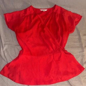 Röd omlottblus från Fórmula - Snygg röd omlottblus från Fórmula med korta ärmar och knyt i sidan. Perfekt för både vardag och fest. Blusen har en v-ringad hals och är tillverkad i ett lätt, mjukt tyg som ger en skön känsla. ❤️